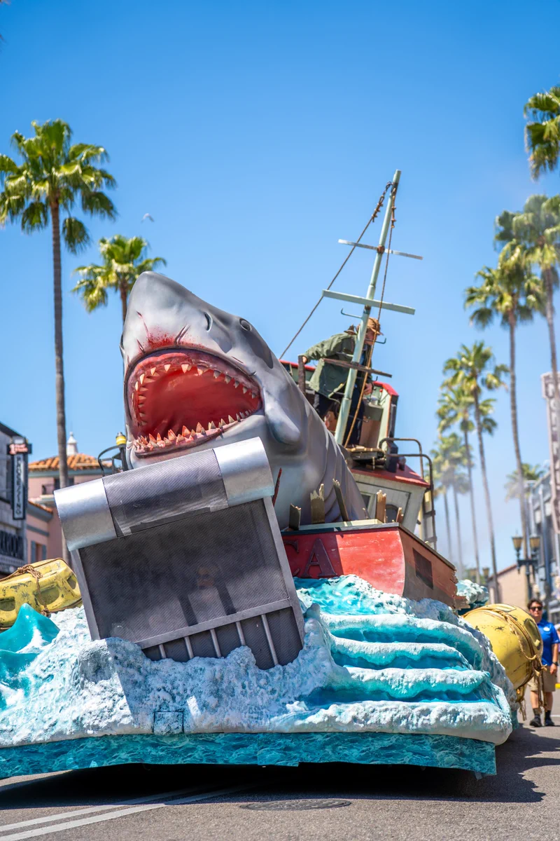 The JAWS float at the Universal Mega Movie Parade. It portrays a gray shark with a wide-open mouth over at a red boat and wave.