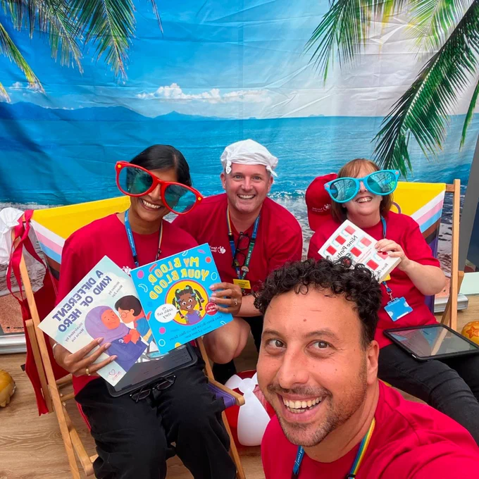 Four smiling adults in red shirts wearing oversized sunglasses, holding children's books and sitting in front of a tropical beach backdrop with palm trees.