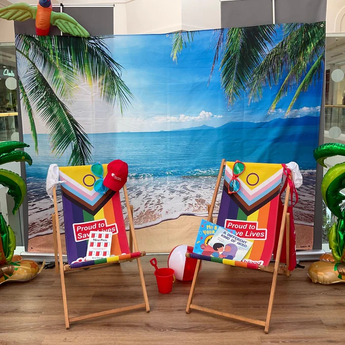 Two colorful beach chairs with 'Proud to Save Lives' signs set against a tropical beach backdrop with palm trees and ocean waves.