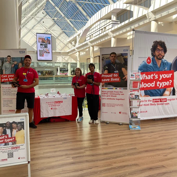 Four volunteers in red shirts stand at an NHS blood donation awareness booth inside a bright shopping mall.
