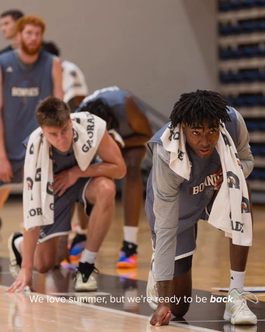 Two basketball players in gray uniforms crouch on the court, focused and ready to start a drill or game.
