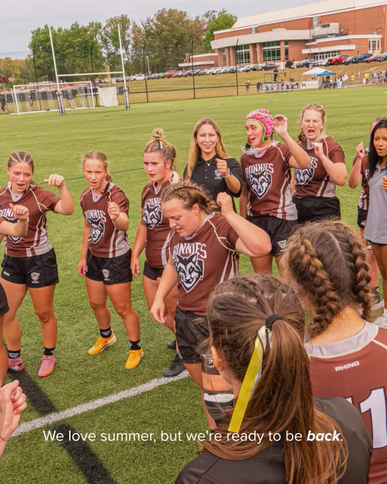 Female rugby players in brown jerseys gather on a green field, showing determination and team spirit before a game.