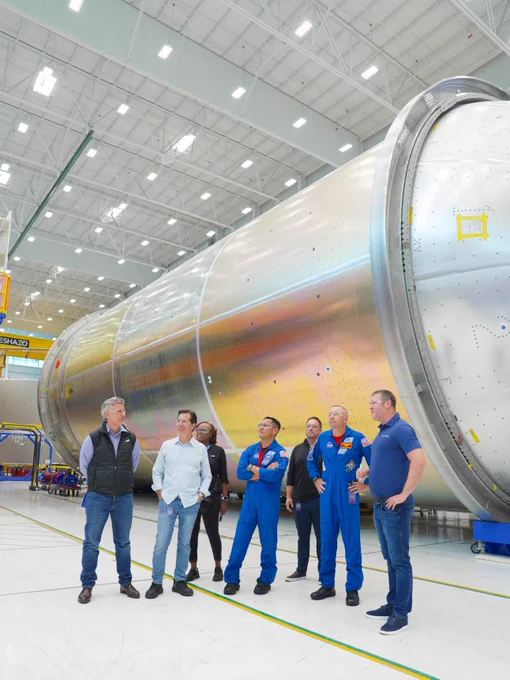 A group of people walking inside a spacious industrial facility with large rocket vehicle components. Two astronauts wearing blue suits are surrounded by others. Bright lighting and large cylindrical structures dominate the background.