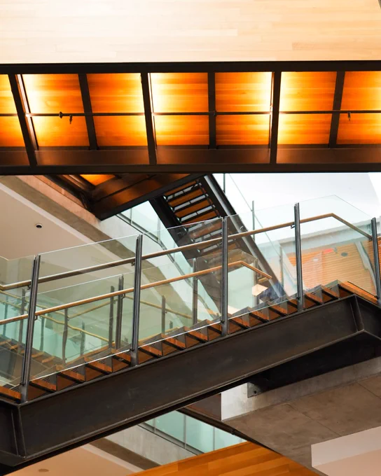 A modern interior features a black metal staircase with wooden treads and glass railings. Warm orange lights illuminate wooden panels on the ceiling above the stairs. A light wooden surface is visible at the top of the frame.