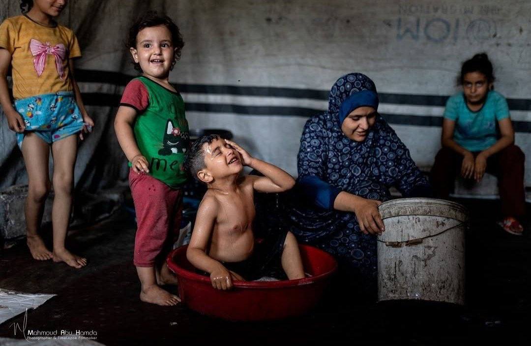 QudsNen's tweet photo. In war-torn Gaza, a mother tries to ease the intense summer heat for her children in their tent, using the small amount of water available to cool their bodies amid soaring temperatures. https://t.co/qH6jWREJqr