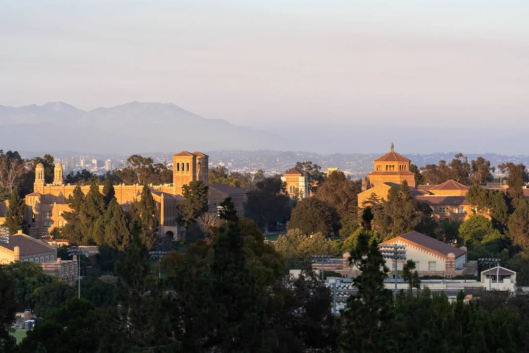 Panoramic view of the UCLA campus with several historic buildings among lush greenery. In the background, the distant mountains are visible under a hazy sky.
