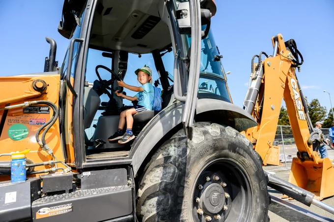 A child sits inside the cab of a large CASE construction vehicle, wearing a light green hat and holding the steering wheel. The vehicle is parked in an outdoor setting under a clear blue sky.