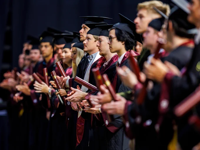 A group of graduates in caps and gowns stand in a line, holding diplomas. They are in an indoor venue, clapping, and appear attentive during a ceremony.