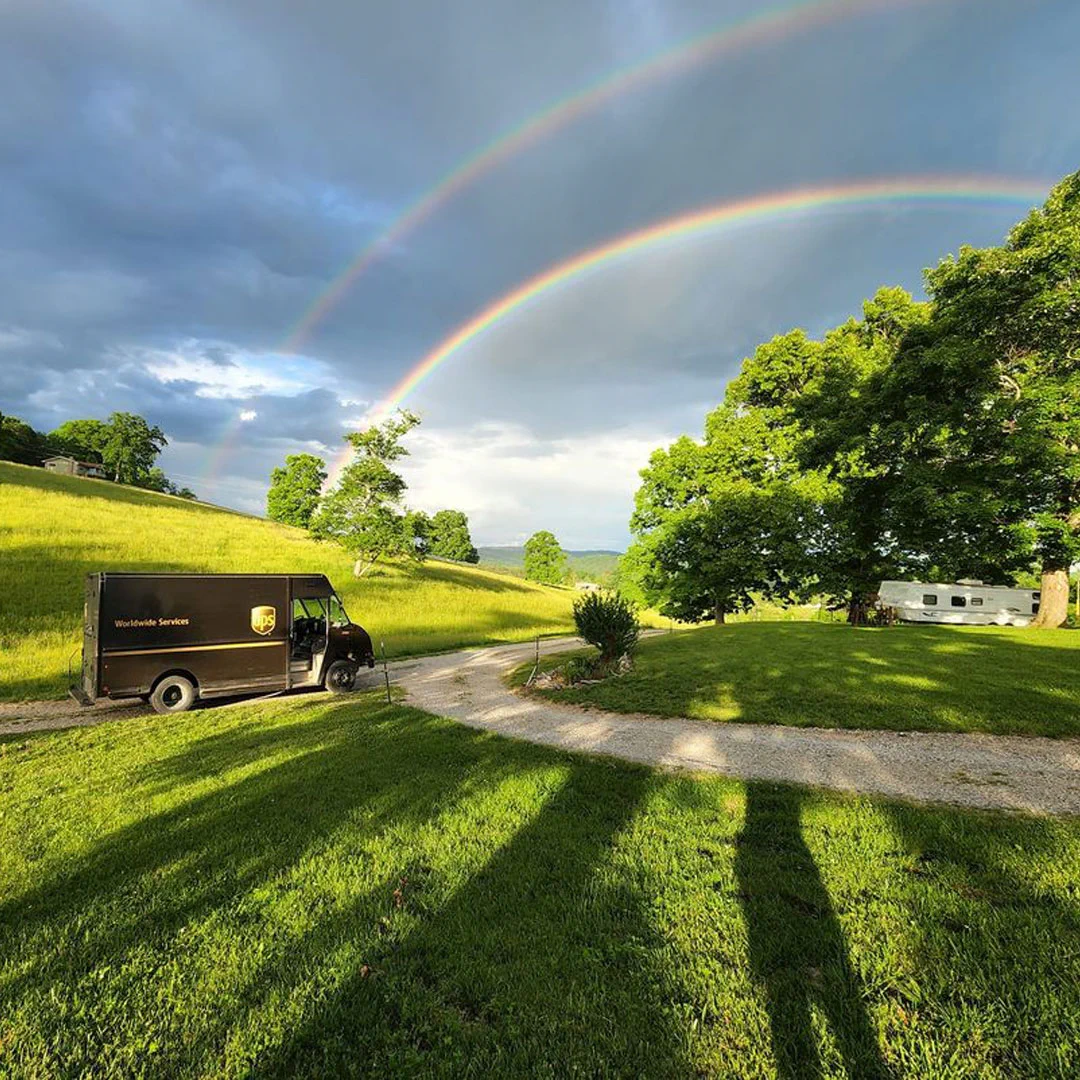 A UPS truck on a gravel path in a grassy landscape with two rainbows in the sky.