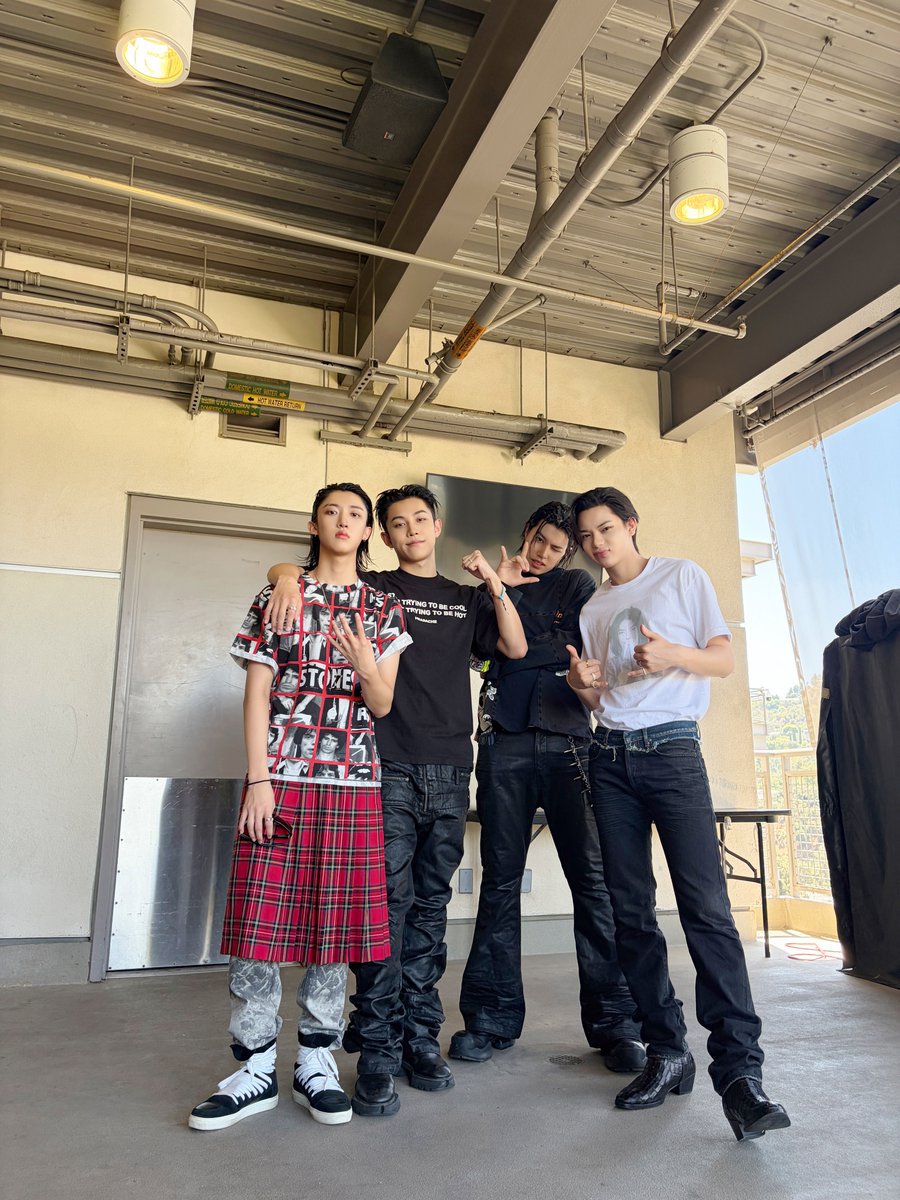 Jidho's tweet photo. four silhouettes standing quiet against the wide open sky of a festival morning. dark denim, white tees catching the last golden hour before the stage lights wake up. hands raised in half-smiles and secret signs, the kind that only happen when the day still feels like a secret. https://t.co/V5pdguYDPO
