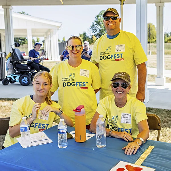 A group of four people are seated and standing at a table during a "Canine Companions DogFest" event. They are all wearing yellow event T-shirts and name tags. Water bottles, a folder, and informational papers are on the table. A person on a mobility scooter is in the background.