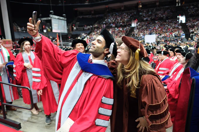 Two individuals in academic regalia take a selfie at a graduation ceremony. They are surrounded by other graduates in robes. The event is taking place in a large indoor arena with many attendees in the background.