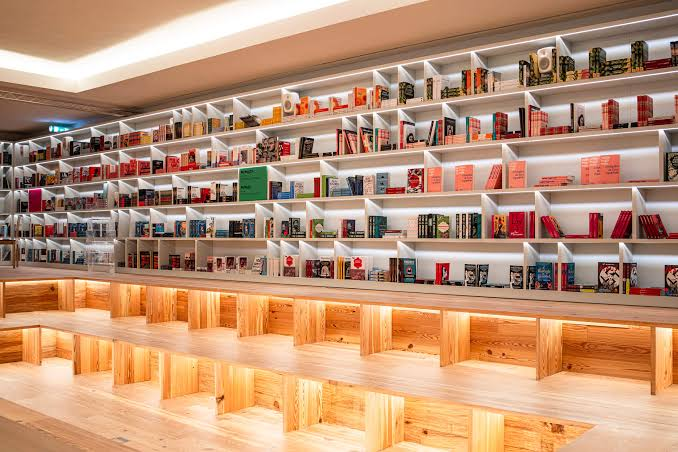 architectizm's tweet photo. デュア・リパが、ポルトガルに「禁書の図書館」をつくったらしい。

2023年から続ける彼女のブッククラブ、Service95 Book Clubと、ポルトの老舗書店Livraria Lelloの一部に開いた「Manifesto Library」。（ちなみにこの書店は世界一美しい書店としても有名）

学校のカリキュラムから外された本、LGBTQIA+を扱うため展示を制限された本など、通常の図書館には起きにくい100冊を収めた常設の図書館。

面白いのは、単なるDuaの選書というより、オンラインのブッククラブを物理的な場所にしたこと。

「検閲」は昔のように焚書や発禁だけでなく、棚におかない、学校で扱わない、なども検閲の一つ。そうやって本と読者が偶然出会う可能性を消していく。

だから対抗策も批判の声明というよりも、もう一度棚に置くこと。この図書館は本を保管するだけでなく、読書、討論、公共的な思考の場として運営されるとのこと。

SNSやニュースレターが本を「届ける」メディアなら、図書館は本を「残し、出会わせ、話させる」物理的な場所。