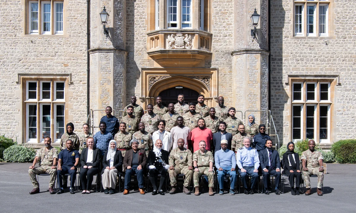Large group of military personnel and civilians posing in front of a historic stone building with arched entrance and windows.