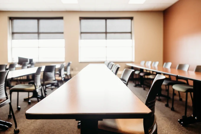 A well-lit, empty classroom with long tables and chairs arranged in rows. Large windows line one side of the room, allowing natural light to pour in.