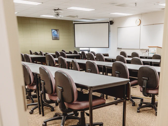 A classroom with rows of empty desks and chairs facing a whiteboard and a lowered projector screen. A clock is visible on the wall.