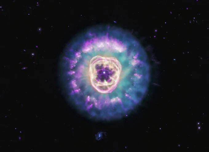 The Lion Nebula, against the black background of space. The nebula is in the center, circular in shape, and looks like a male lion’s head. In the very center is a small, bluish-white circle with eight-point diffraction spikes, a white dwarf star. Surrounding the star are light purple-red cavernous bubbles and shell-like rings. The bubbles and shells collectively form an oval with two small, wide arcs near the top, reminiscent of a lion’s face and its ears. What appears to be extended outward from the lion’s face is a thick ring of cyan material. The width of the ring is consistent throughout and resembles a mane. The area closest to the lion’s face looks cloudy, while the edges of the mane look like there are purple clumps of dust with comet-like tails and strands. In the background are distant galaxies and stars. Some galaxies appear as small purple points of light and others with visible blue spiral structures. A couple of the stars have diffraction spikes.