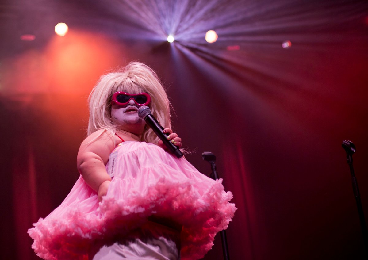 Tamm Reynolds, Midgitte Bardot’s Shooting From Below. Photo by Holly Revell
Performer Tamm Reynolds on stage in drag. They wear a big blond wig, a tulle frilly pink top, red sunglasses and hold a microphone.