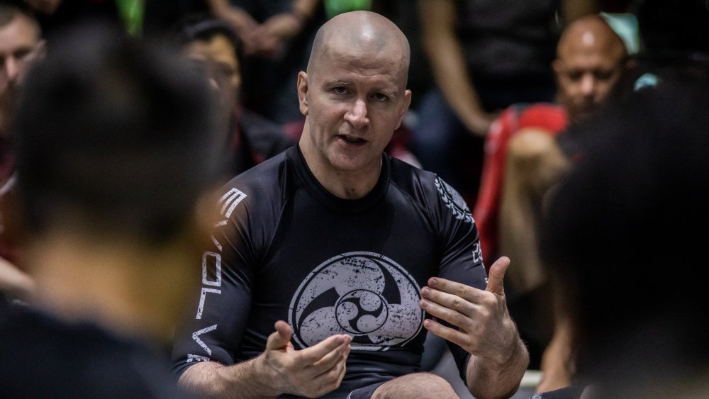 rfatsta's tweet photo. BJJ'in Einstein'i: JOHN DANAHER

John Danaher, modern Brazilian Jiu-Jitsu ve submission grappling tarihinin en etkili antrenörlerinden biri. İlginç olan şu: Danaher aslında klasik anlamda büyük bir yarışmacı değil; felsefe eğitimi almış, akademik düşünce yapısına sahip bir öğretmen olarak jiu-jitsu'ya girdi ve zamanla Gordon Ryan, Garry Tonon, Craig Jones, Nicky Ryan, Eddie Cummings ve Georges St-Pierre gibi isimlerin gelişiminde rol oynadı. 

Onu özel yapan şey yalnızca iyi teknikler öğretmesi değil. Jiu-jitsu'yu bir "teknikler koleksiyonu" olmaktan çıkarıp birbirine bağlı sistemler bütünü olarak düşünmesi. Özellikle leg-lock, back attack ve submission sistemlerinin modern grappling'e yerleşmesinde çok büyük etkisi oldu.

1. John Danaher kimdir?

John Bernard Danaher, 1967'de Washington D.C.'de doğdu ve Yeni Zelanda'da büyüdü. Columbia University'de felsefe eğitimi aldı; özellikle felsefe ve akademik düşünce dünyasında ilerledi. Ancak üniversite yıllarında geçimini sağlamak için New York'ta güvenlik görevlisi/bouncer olarak da çalışıyordu. 

Jiu-jitsu'ya oldukça geç başladı.

Yaklaşık 28 yaşındaydı.

Bu önemli çünkü BJJ'de birçok üst düzey sporcu çocukluk veya ergenlik döneminde başlarken Danaher yetişkin olarak başladı. Kendi anlatımına göre ilk dönemlerinde dövüş sanatlarına karşı oldukça farklı bir bakış açısı vardı ve jiu-jitsu ile tanışması biraz tesadüfi gerçekleşti. 

Onun hayatındaki dönüm noktası:

Renzo Gracie.
Danaher, New York'taki Renzo Gracie akademisine girdi ve Renzo'nun öğrencisi oldu.

Danaher, beyaz kuşaktan siyah kuşağa kadar Renzo'nun öğrencisi olarak kaldı ve kendi anlatımına göre hiçbir zaman başka bir hocaya geçmedi. 

Bu ilişki onun bütün kariyerinin temelini oluşturdu.

2. Danaher neden yarışmacı olarak ünlü olmadı?

Burada önemli bir ayrıntı var.

Danaher'in profesyonel yarışma kariyeri yok denecek kadar sınırlı.

Bunun önemli sebeplerinden biri ciddi kalça ve bacak problemleri yaşamasıydı. Bu nedenle kendisini aktif bir yarışmacı olarak değil, daha çok öğretmen ve araştırmacı olarak geliştirdi. 

Bu yüzden Danaher'in hikâyesi:

"Harika bir şampiyon oldu, sonra antrenörlüğe geçti."

hikâyesi değil.

Tam tersine:

"Kendi vücudu yarışmacı olmasına izin vermedi ve bunun yerine dövüş sanatını analiz etmeye, öğretmeye ve sistemleştirmeye yöneldi."

Bu da onun en büyük avantajlarından birine dönüştü.

3. Felsefe eğitimi neden bu kadar önemli?

Bence Danaher'i anlamanın anahtarı burası.

Danaher jiu-jitsu'yu genellikle şöyle düşünmüyor:

"Şu pozisyonda şu hareketi yap."

Onun yaklaşımı daha çok:

"Bu pozisyon neden oluştu? Rakibin amacı ne? Benim amacım ne? Rakibin hangi reaksiyonları mümkün? Her reaksiyona karşı hangi cevabı verebilirim?"

şeklinde.

Kendi röportajlarında akademik geçmişinin öğretme biçimini ciddi şekilde etkilediğini açıkça anlatıyor. Akademik ortamda karmaşık fikirleri parçalara ayırarak öğretmesinin jiu-jitsu eğitimine doğrudan geçtiğini söylüyor. 

Bu yüzden Danaher'in dersleri bazen BJJ dersinden çok felsefe/mühendislik dersi gibi algılanabiliyor.

4. Danaher'in jiu-jitsu anlayışı

Danaher'in en önemli fikri:

"Jiu-jitsu bir sistemdir."
Örneğin klasik BJJ'yi kabaca şöyle sıralıyor:

Rakibi yere indir.

Guard'ını geç.

Dominant pozisyon elde et.

Submission yap.

Bu sistemde hareketler birbirine bağlıdır.

Danaher'in yaptığı şey ise:

Bu büyük sistemi daha küçük sistemlere ayırmak.

Örneğin:

Leg-lock sistemi

Back attack sistemi

Front-headlock sistemi

Kimura sistemi

Armbar sistemi

Triangle sistemi

Takedown sistemi

Pinning/control sistemi

Sonra bunları birbirleriyle bağlantılı hale getirmek.

Yani:

System → Subsystem → Connection → Reaction → Counter

mantığı.

Danaher bunu yıllar içerisinde özellikle öğrencileri üzerinde test etti. 

⬇️