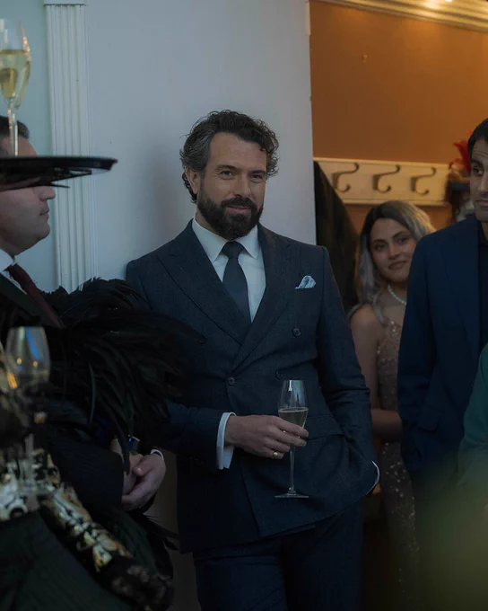 A bearded man (Tom Cullen) in a dark double-breasted suit holds a glass of sparkling wine while standing among guests at a formal party.