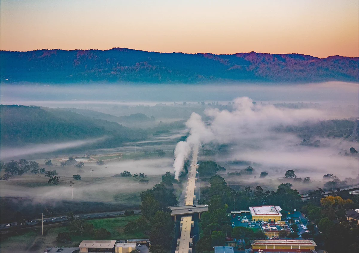 A scenic aerial view of a long, straight path of white steam rising through a misty valley at sunrise. Foothills and mountains form the background, while buildings and greenery are in the foreground, including part of the SLAC National Accelerator Laboratory. Credit: Olivier Bonin/SLAC