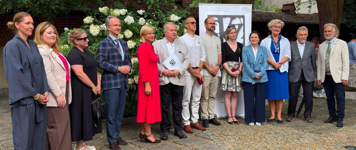 Grupowe zdjęcie uczestników wydarzenia w plenerze. Jedenaście osób pozuje przed budynkiem, na tle zieleni i kwitnących hortensji. Wśród uczestników znajduje się wiceministra kultury i dziedzictwa narodowego Bożena Żelazowska. Za grupą widoczny jest baner wydarzenia.