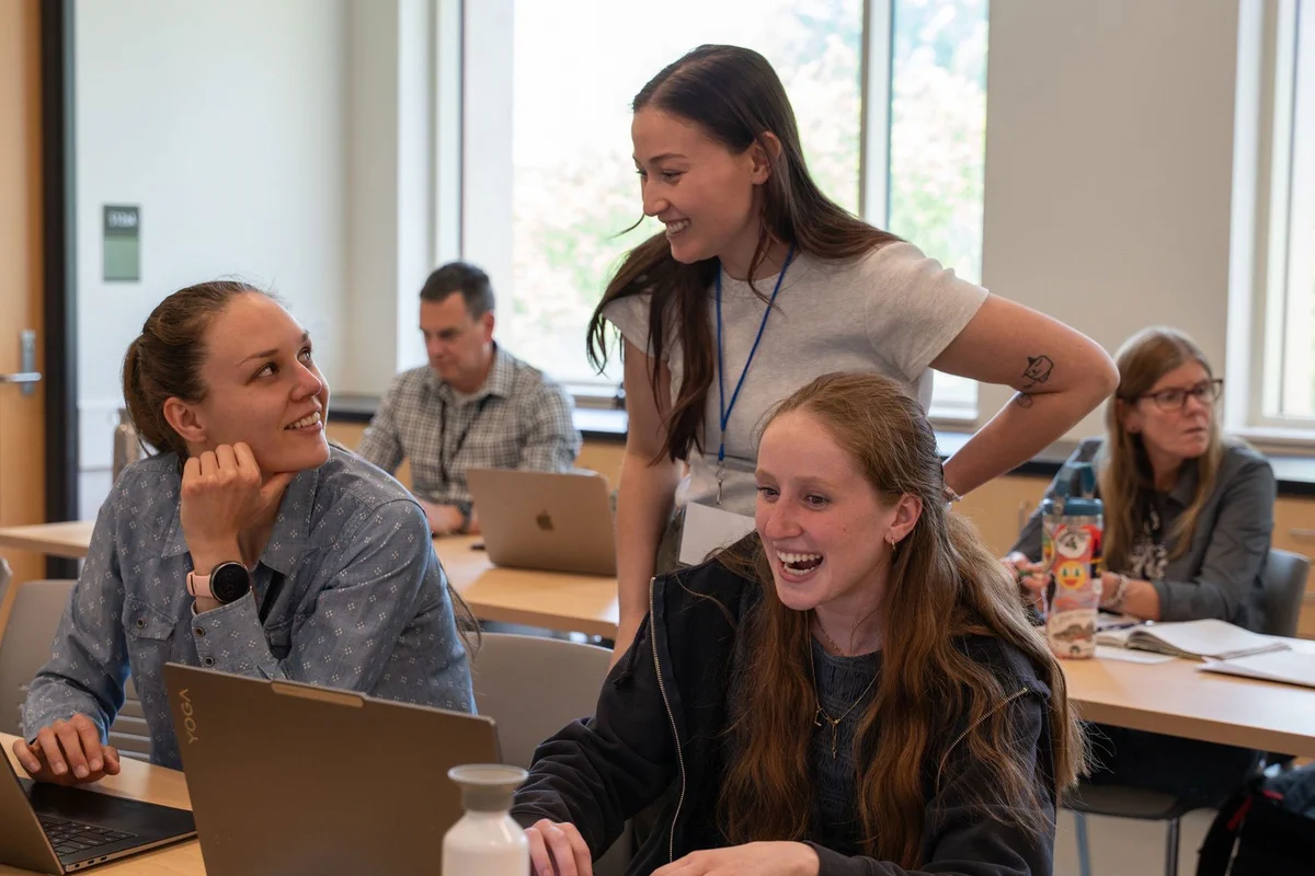 Three people smiling and in discussion looking at a laptop in a room of other people working on laptops.