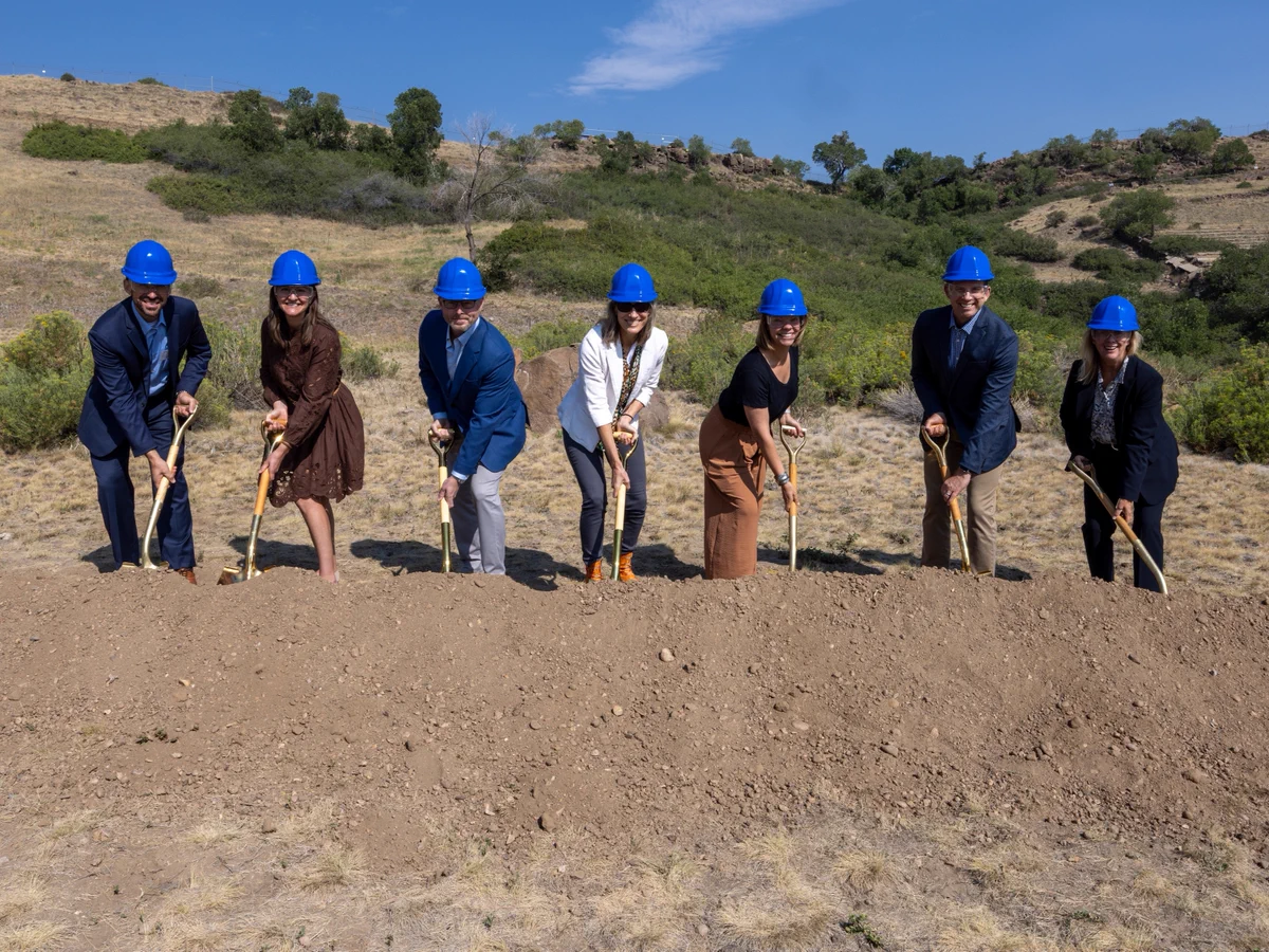 A group of seven people wearing blue hard hats are participating in a groundbreaking ceremony outdoors. They stand in a line, each holding a shovel ready to dig into the dirt. The background features a hilly landscape with grass and shrubs under a blue sky.