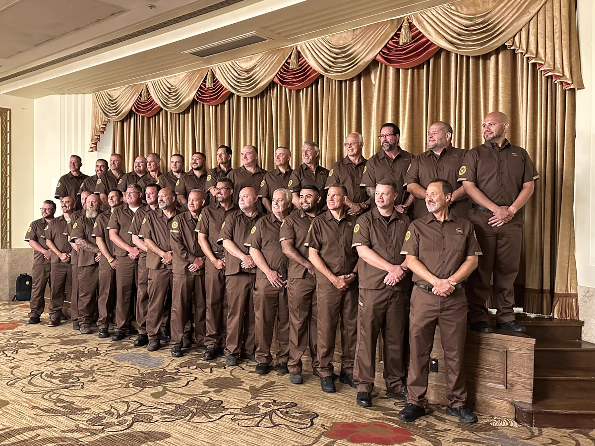 A group of drivers in uniforms posing together in front of gold and red drapery.