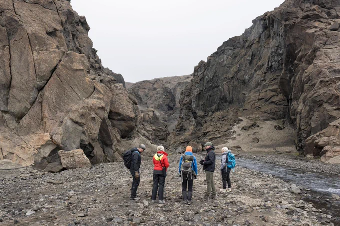 A small group of people wearing outdoor gear stand on a rocky valley floor in Iceland, surrounded by steep, jagged cliffs of dark volcanic rock. They appear to be discussing the terrain while a narrow stream flows beside them. The landscape is rugged and barren, with no vegetation visible. Credit: NASA/James Blair