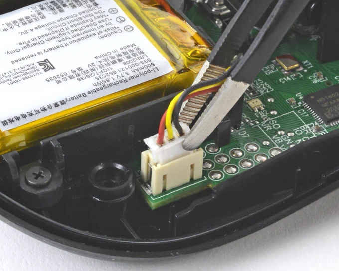 Tweezers disconnect a red, yellow, and black wire connector from a green circuit board next to a battery inside a disassembled mouse.