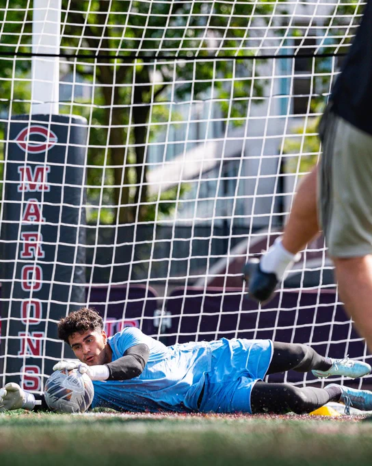 A male soccer goalkeeper in a light blue kit and black undersleeves lies on green turf, hands on a soccer ball. He is in front of a goal net. Behind the net is a dark banner with 'MARONS' and a 'C' logo. Another player's leg is blurred in the upper right.