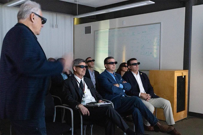 A group of people is sitting in a dimly lit room wearing dark glasses, facing a speaker. A whiteboard with diagrams is visible in the background. Credit: Sam Soon/SLAC
