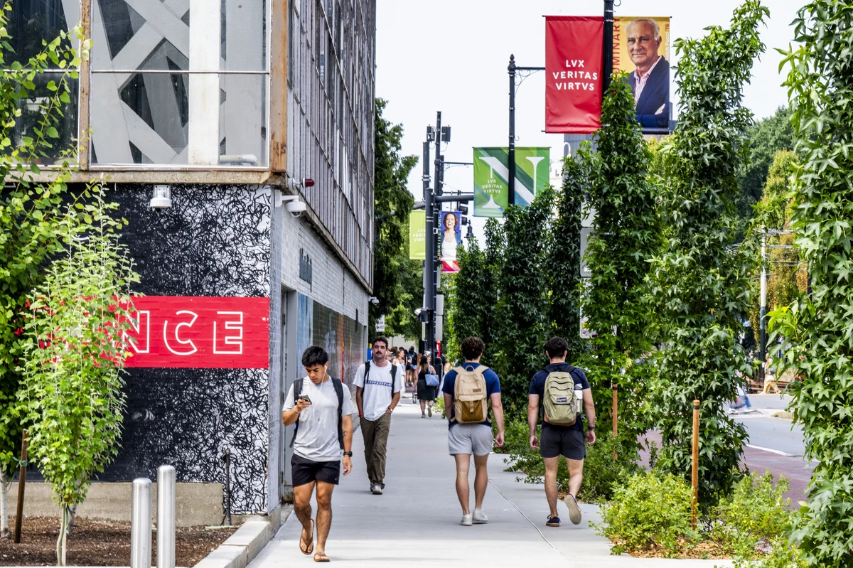 Young adults walk on a tree-lined city sidewalk. A man in the foreground looks at his phone. Buildings with textured walls frame the left. Lampposts display red and green banners with 'LVX VERITAS VIRTUS' and portraits.