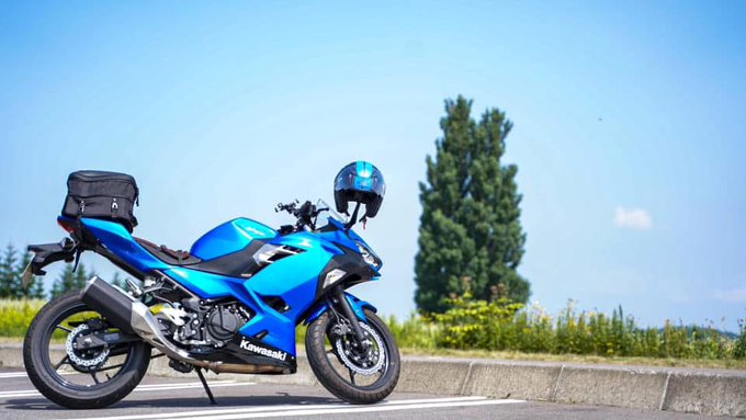 Side profile photograph of a blue Kawasaki motorcycle parked on the side of a road with a black helmet on the handlebars and a tail bag attached, set against a background of trees and sky.
