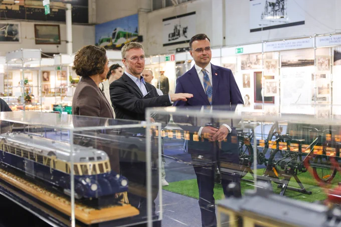 Wiceminister kultury i dziedzictwa narodowego Maciej Wróbel zwiedza ekspozycję w Stacji Muzeum w Warszawie w towarzystwie przedstawicieli muzeum. Grupa stoi przy gablotach z modelami historycznych pojazdów kolejowych. Jeden z mężczyzn wskazuje element wystawy i opowiada o eksponatach.