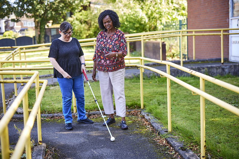 Two individuals are outdoors on a pathway with yellow railings.