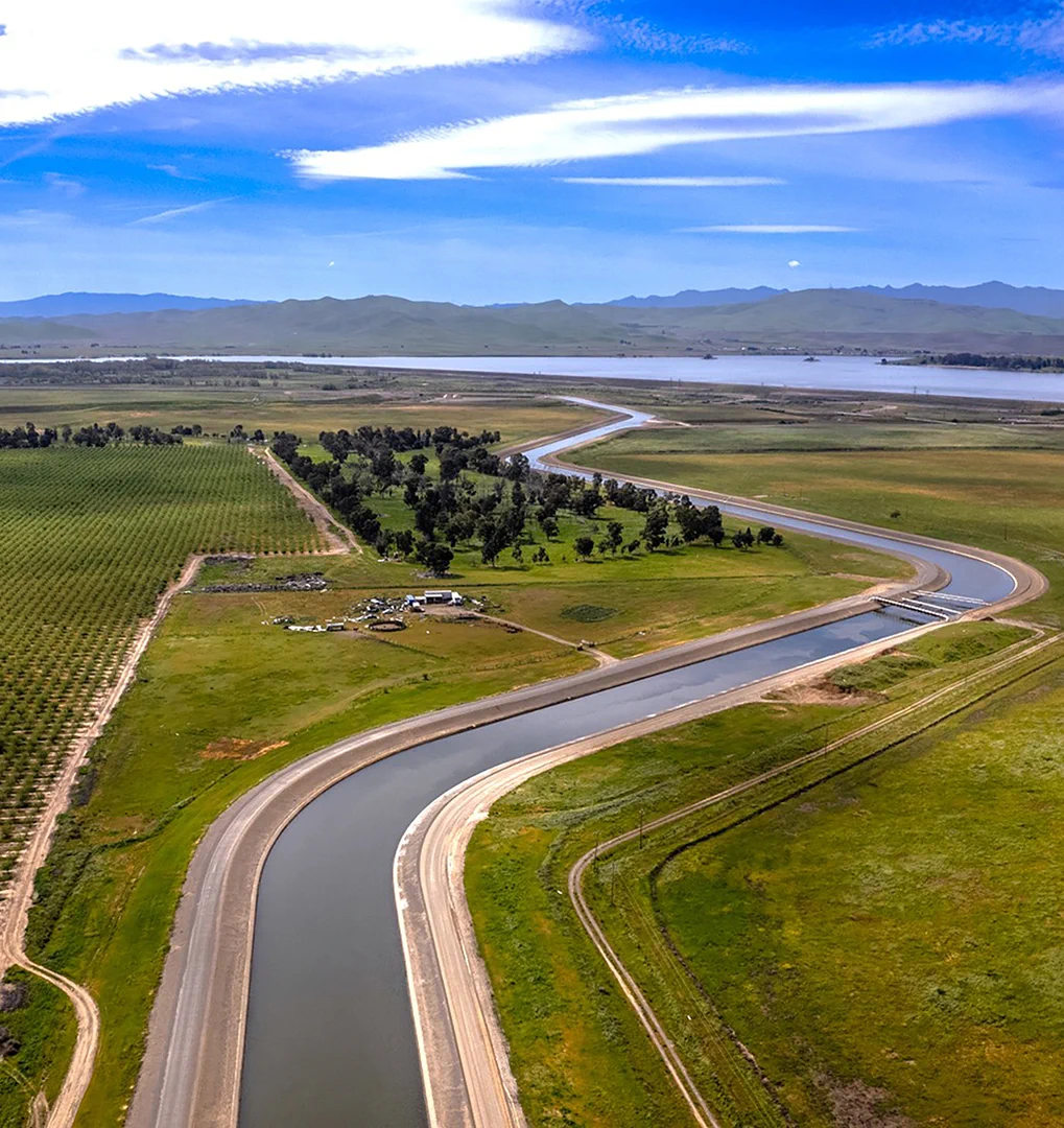 A winding irrigation canal cuts through green farmland under a bright blue sky with scattered clouds.