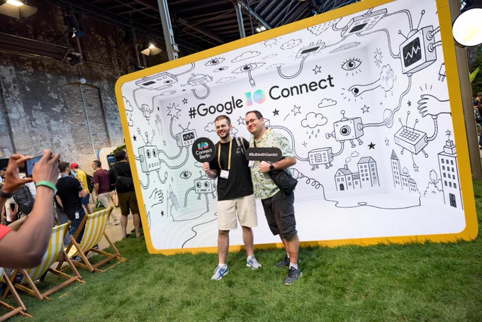 Two people  smile with props in hand in front of a photo backdrop with text "#Google I/O Connect" and illustrations related to imaginative tech.