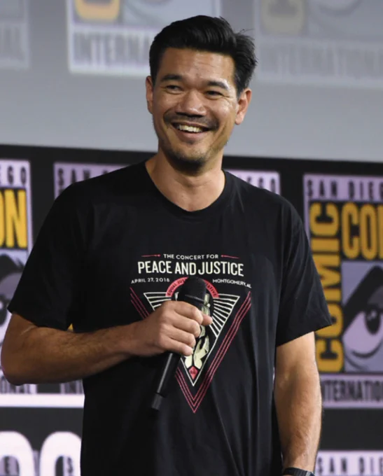 A person holding a microphone is smiling while standing in front of a San Diego Comic-Con background. They are wearing a t-shirt with the text "The Concert for Peace and Justice" and an image of a dove.