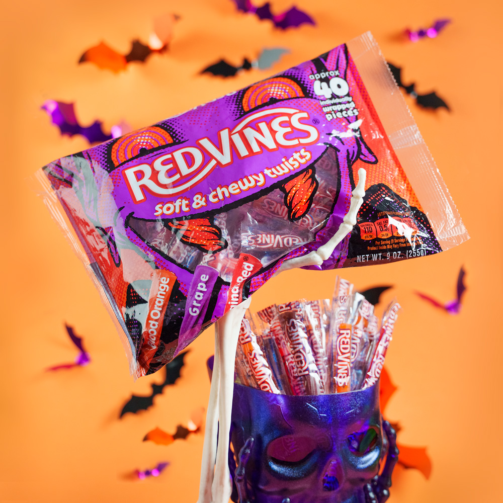 The image shows a package of Red Vines soft and chewy twist candies, labeled as grape and orange flavors. The package is artistically opened, with some candies displayed in a decorative skull-shaped container. The background is orange with bat-shaped decorations.