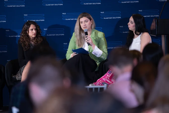 A panel discussion at Yale Planetary Solutions, with three individuals seated on stage. The background features a blue banner with the event's logo. A vase with blue flowers is visible on a small table. The person in the center is speaking into a microphone.