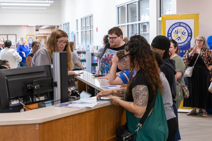 A group of individuals lines up at a service counter in a library setting. Staff assist visitors.
