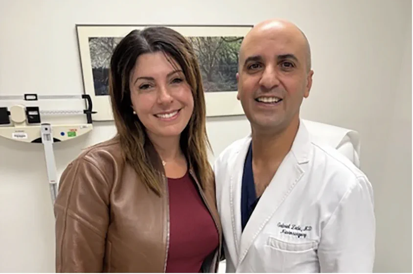 A smiling woman in a brown jacket and burgundy top stands beside a smiling medical professional wearing a white coat and navy scrubs in a clinical exam room. Medical equipment and framed artwork are visible in the background