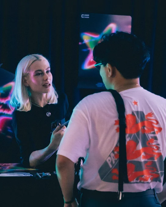 Phoebe Bridgers in a black top holding a pen and speaking across a table to a person whose back is to the camera. Glowing, abstract posters are visible in the dark background, suggesting a fan signing or event.