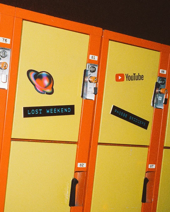 Bright yellow and orange vintage lockers. One locker door features an iridescent planet sticker and a label reading "LOST WEEKEND". The adjacent locker has a YouTube logo sticker and a label reading "PHOEBE BRIDGERS".