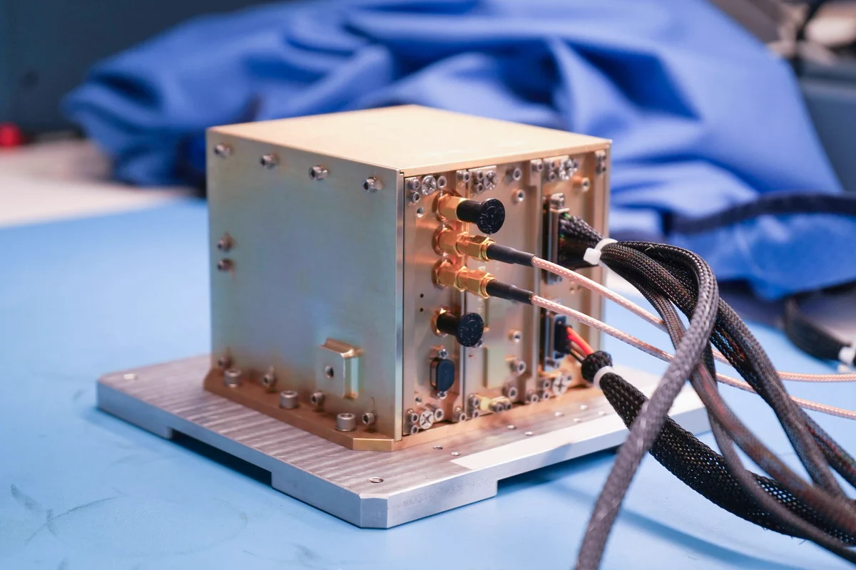 A small, rectangular, metal navigation receiver sits on a silver-colored mounting plate atop a blue laboratory workbench. Several cables connect to ports on the box and extend out of frame. The NavCube3-mini receiver is about half the size of a shoebox and is pictured in a NASA Goddard laboratory before delivery for integration into the Altus-1 lunar relay satellite. Credit: NASA