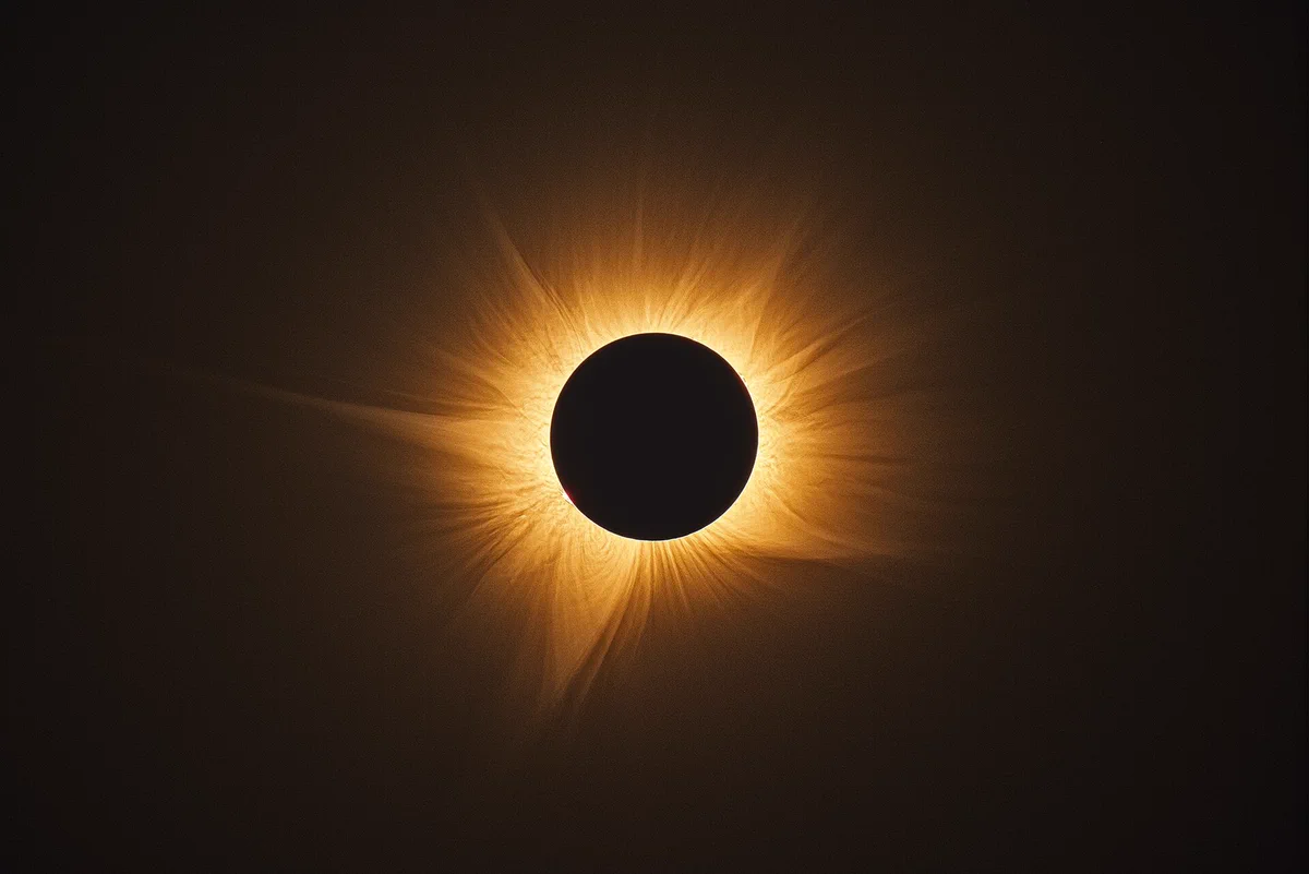 A total solar eclipse seen against a black sky. The Moon completely covers the Sun, forming a dark circular silhouette surrounded by the Sun’s glowing corona, which extends outward in delicate golden streamers of light.