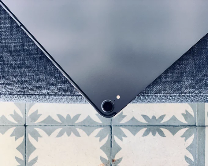 Close-up of the corner of an iPad with its rear camera and flash visible, resting on a woven gray fabric surface above a patterned tile floor.