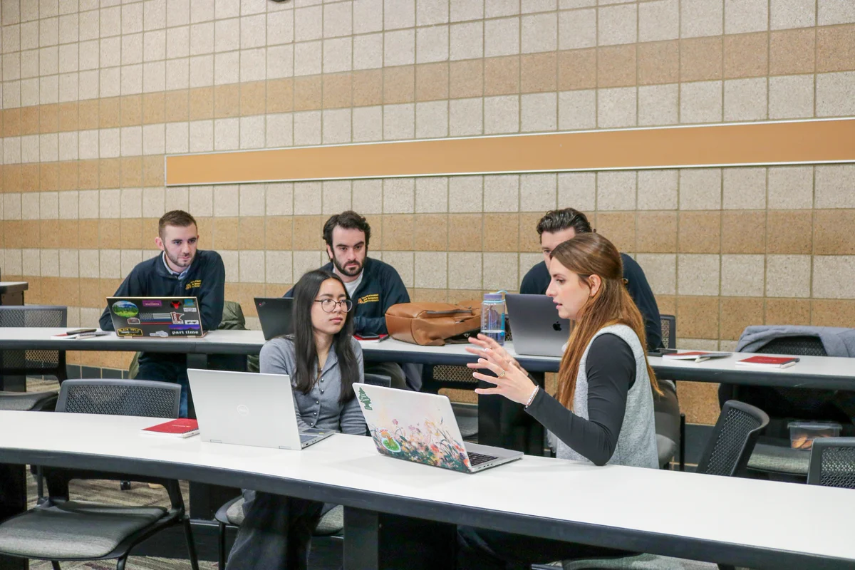 A group of people sitting in a classroom with laptops. They appear to be engaged in discussion. The setting includes long tables and beige tiled walls.